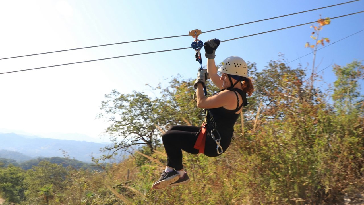 A zipliner gliding above the forest canopy, with the hills around Chiang Mai behind