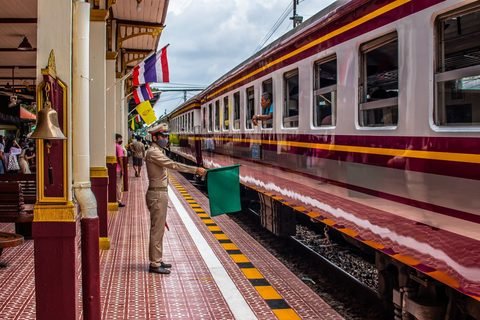 A station officer in uniform holds out a green flag beside a passenger train at Ayutthaya railway station