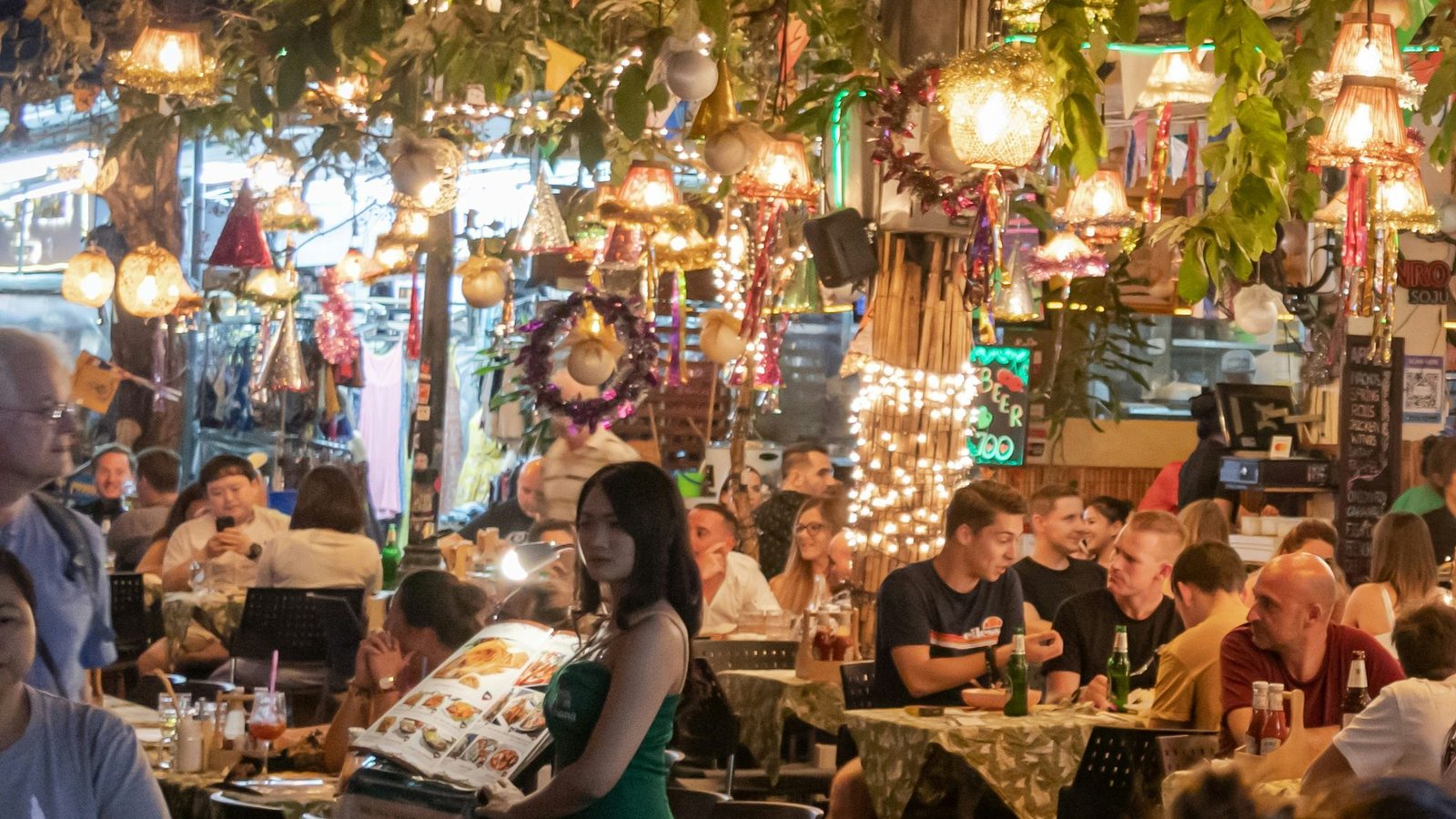 An open-air bar busy with people at night, under string lights, hanging lamps and rows of coloured bunting