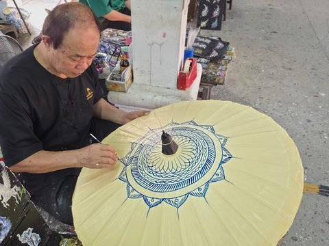Une personne en chemise noire peint au pinceau fin un mandala bleu sur une ombrelle jaune pâle