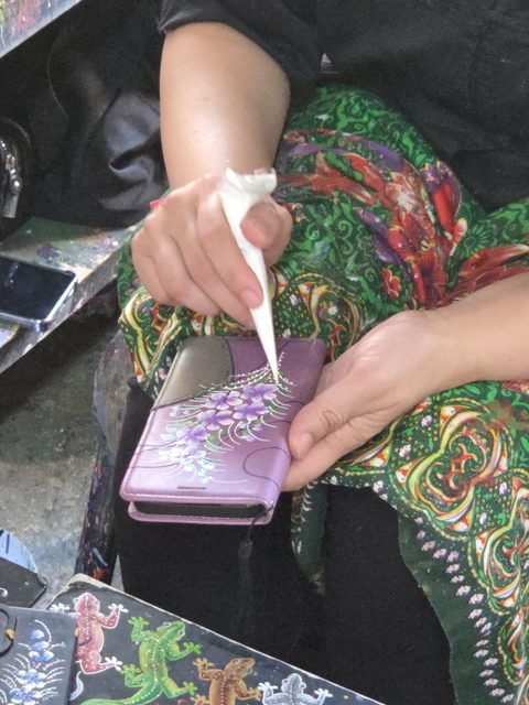 Une main dépose de la peinture blanche au cornet de papier sur un étui de téléphone lilas déjà peint de fleurs