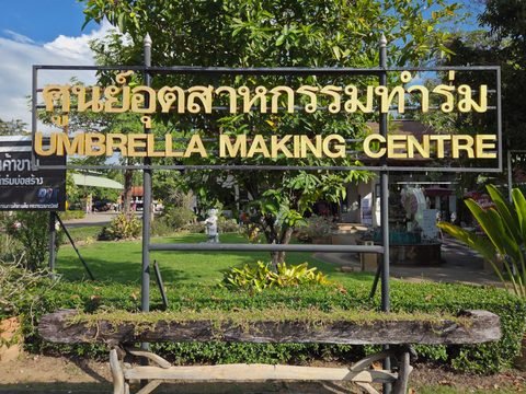 Une enseigne dorée « Umbrella Making Centre », en thaï et en anglais, au-dessus d'une pelouse, avec une fontaine et des bâtiments à l'arrière