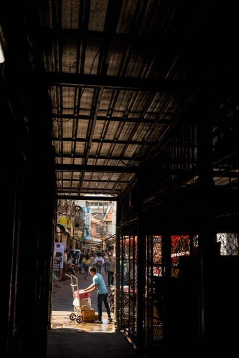 A covered market passage opening onto a busy daylit street, where shoppers carry bags and a man pushes a hand trolley