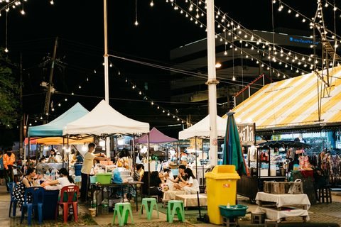 A night market under strings of lights, with food stalls, low tables and plastic stools where people sit and eat