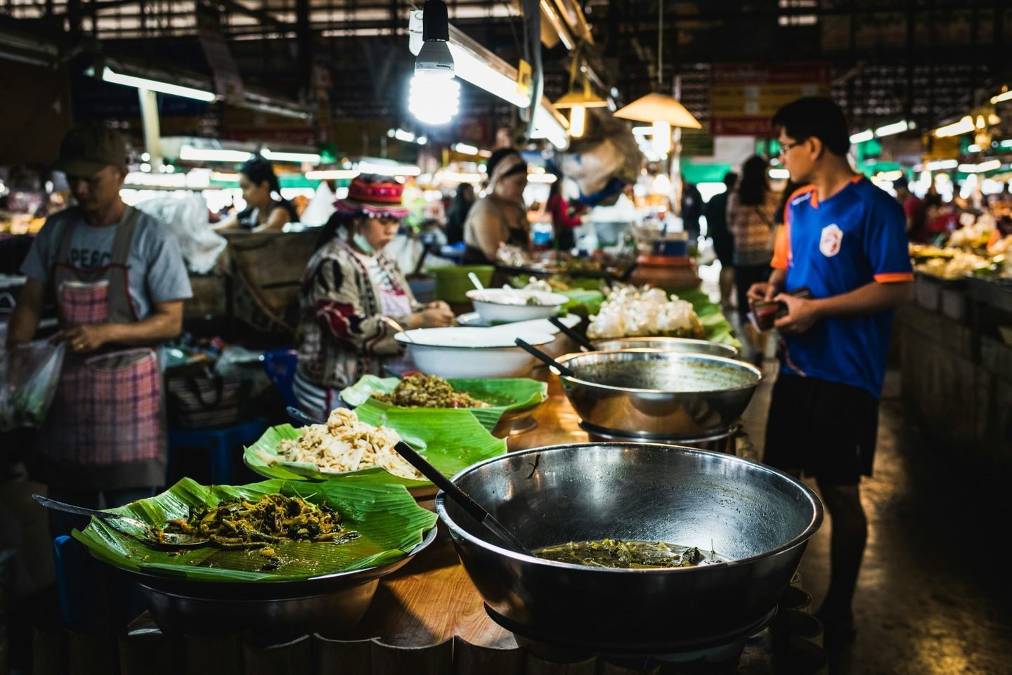 Prepared dishes on banana leaves and in wide pans on the counter of a covered market under fluorescent lights
