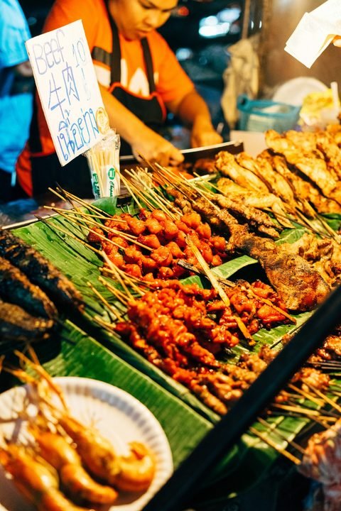 Skewers and grilled fish laid out on banana leaves at an evening stall, with a handwritten sign in English, Chinese and Thai