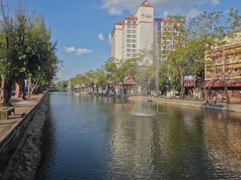 Chiang Mai's east moat looking north from beside Tha Phae Gate square: a fountain in the water, trees along both banks and a tall white hotel on the outer side