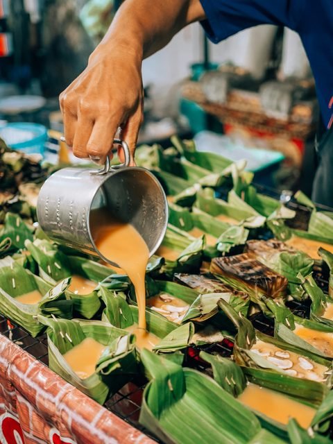 A vendor pouring a mixture into banana-leaf cups over a grill at a market stall