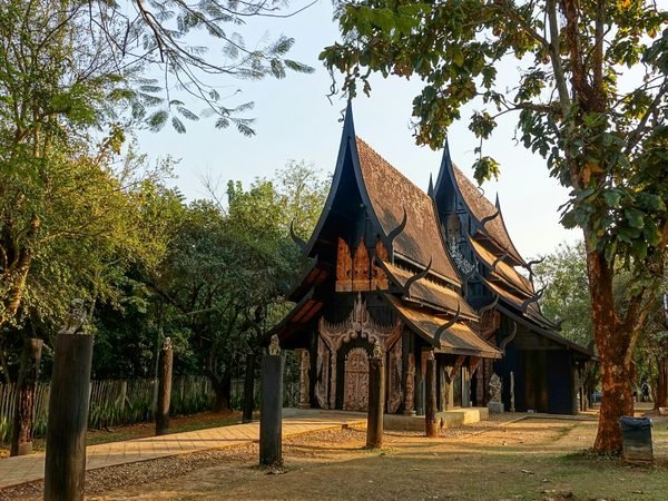 A dark wooden pavilion with steep roofs and horn-shaped finials among the trees at Baan Dam