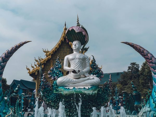 A white seated figure holding an alms bowl above a fountain, framed by two curved blue sculptures, in front of the blue and gold hall of Wat Rong Suea Ten