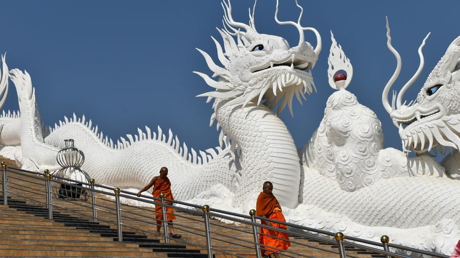 Two monks in orange robes on the stairway at Wat Huay Pla Kang, flanked by giant white dragons against a deep blue sky