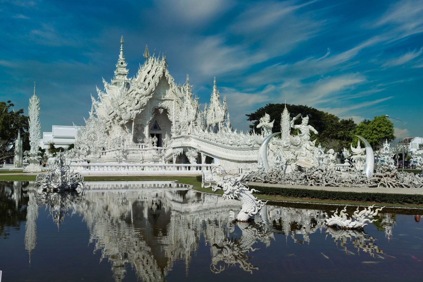 Wat Rong Khun, the White Temple: its ornate white ordination hall and sculptures reflected in the pond in front of it