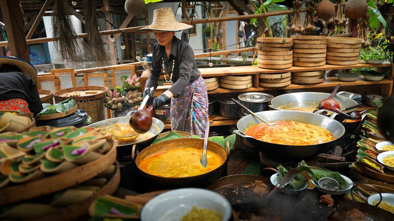 A cook in a straw hat stirring wide pans of curry beside stacked bamboo steamers at a northern Thai kitchen