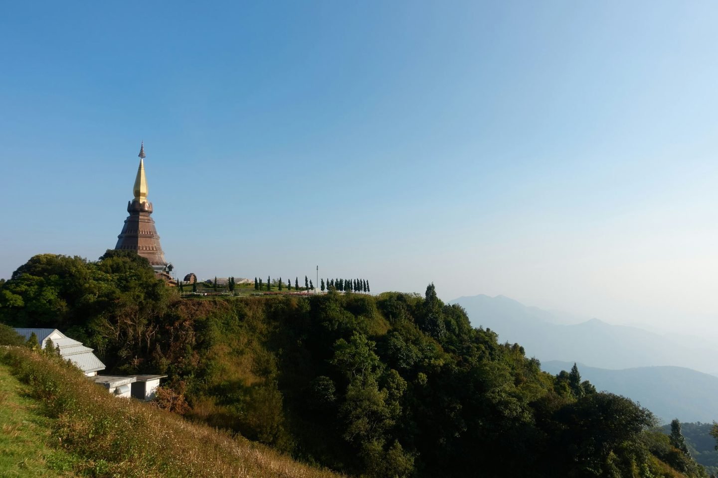 A brown and gold chedi standing on a wooded mountain shoulder, ranges of hills fading into haze behind it