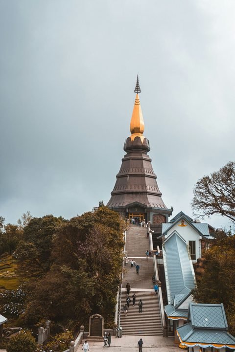A long flight of steps climbing to a brown chedi with a gold spire, visitors going up under a grey sky