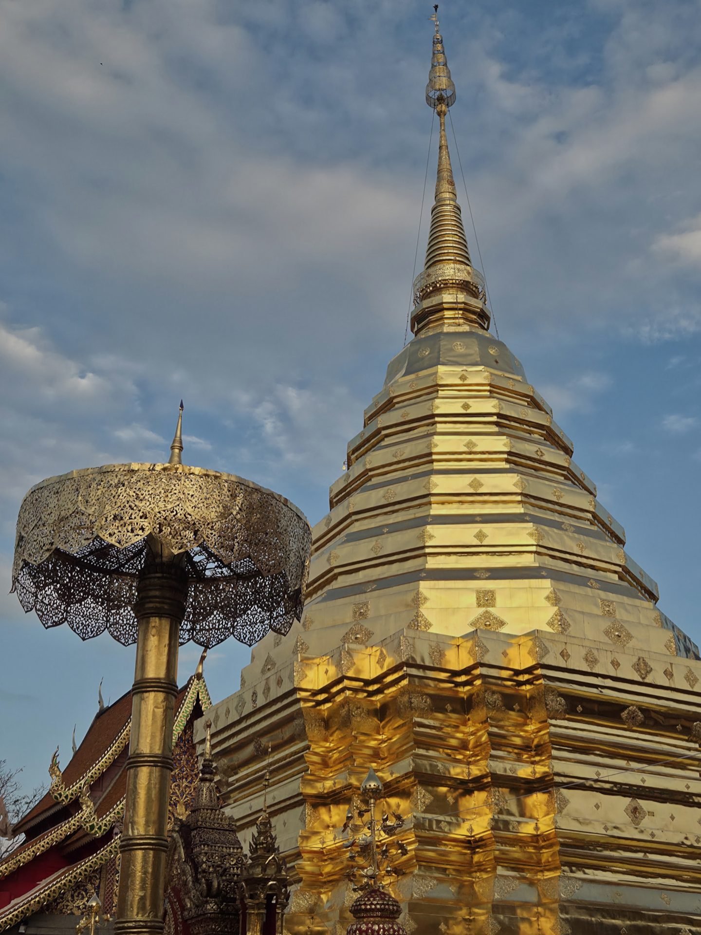 Le chedi doré du Wat Phra That Doi Suthep sous un ciel bleu nuageux, le soleil bas éclairant sa base, et devant lui un parasol doré ajouré au sommet d'un haut mât