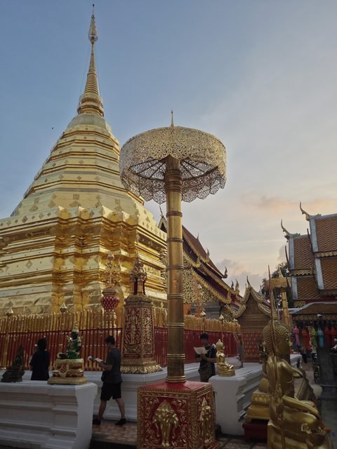 Le chedi doré dans la lumière de fin d'après-midi, un parasol d'angle sur son socle rouge, la grille dorée, un Bouddha vert sur un piédestal et, à droite, les toits de tuiles d'une salle