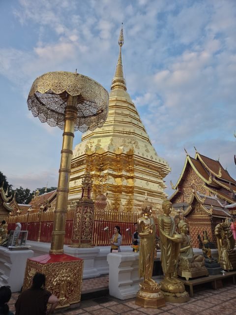 Le chedi doré et l'un des grands parasols dorés qui l'entourent, derrière la grille, des statues dorées debout au premier plan, et deux personnes qui font le tour, des fleurs à la main