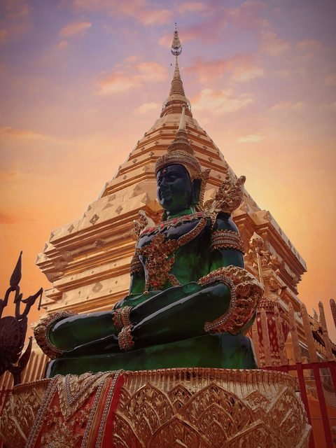 A green Buddha image seated in front of the gilded chedi of Wat Phra That Doi Suthep, against an evening sky