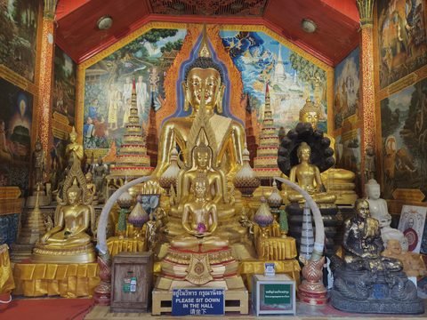Un autel chargé de Bouddhas dorés devant des peintures murales, encadré de deux défenses recourbées, et un panneau en thaï, en anglais et en chinois qui demande de s'asseoir dans la salle