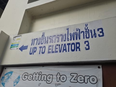 Un panneau peint sur un mur blanc, avec une flèche : en thaï, l'accès au « tram électrique », niveau 3, et en anglais, « Up to elevator 3 »