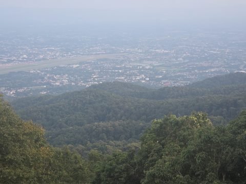 Le panorama depuis le point de vue du temple, dans la brume de fin d'après-midi : des pentes boisées au premier plan, puis la longue piste de l'aéroport de Chiang Mai et la plaine bâtie, qui se perd dans un ciel pâle