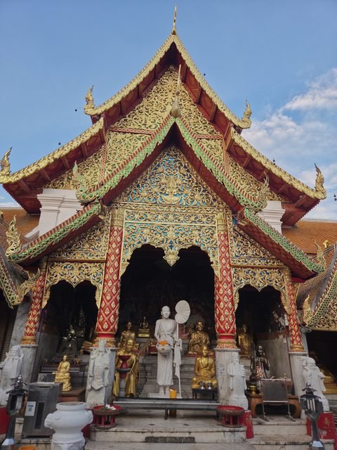 Le porche d'une salle du temple en plein jour : toit de tuiles rouges à plusieurs étages, pignon sculpté et doré sur fond bleu, statue blanche d'un moine debout parmi des figures dorées assises