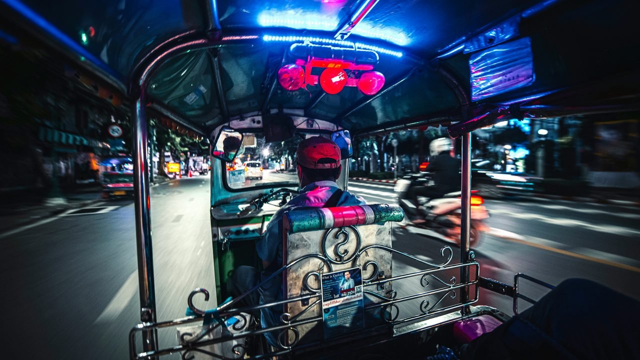 The view from the back of a tuk-tuk at night, neon under the canopy and the traffic blurred past on either side