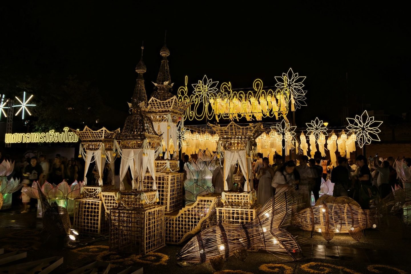 A lantern and bamboo installation lit up at night, with people walking around it and an illuminated Chiang Mai Municipality sign in Thai on the left