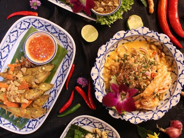 Finished Thai dishes laid out on patterned plates and banana leaf, chillies, shallots, lime and ginger around them