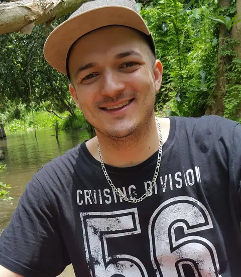Gregory smiling, in a cap and a dark T-shirt, with trees and water behind him