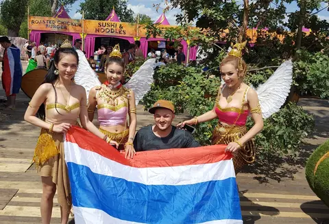 Gregory crouching behind a Thai flag held up by three women in gold costumes with white wings, outdoors at a festival