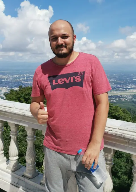 Gregory giving a thumbs-up beside a white balustrade, with a city spread across the plain and clouds behind him