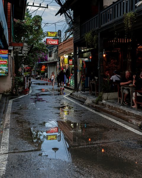 A narrow street of bars and cafes reflected in wet tarmac at dusk