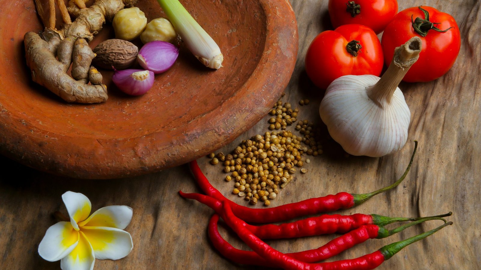 Thai cooking ingredients on a wooden table: galangal and lemongrass in a clay mortar, shallots, garlic, tomatoes, coriander seed and red chillies