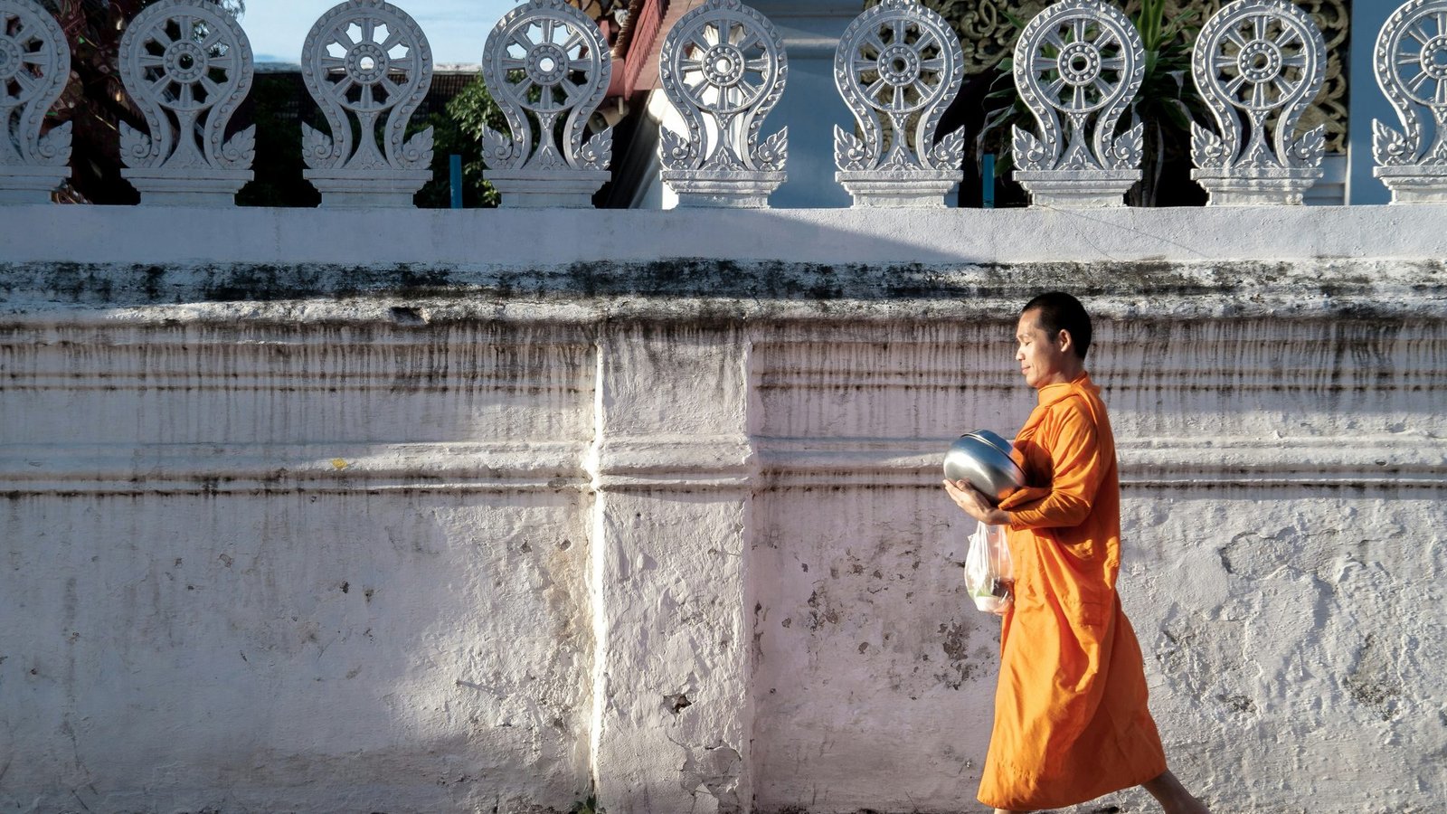 A monk in orange robes walking barefoot past a whitewashed temple wall topped with carved dharma wheels, an alms bowl under his arm