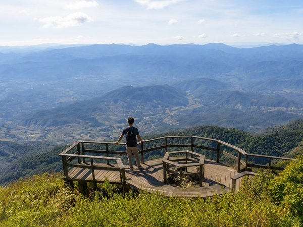 A walker alone on the wooden viewing deck at the end of the Kew Mae Pan ridge, ranges of hills fading behind