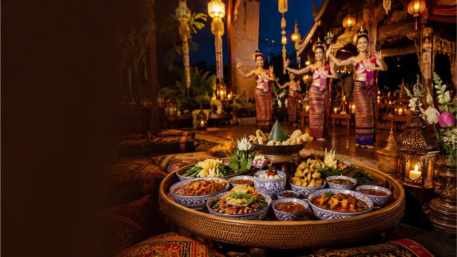 A low round tray set with small bowls of northern Thai dishes, with dancers in Lanna costume performing behind it among lanterns and candles