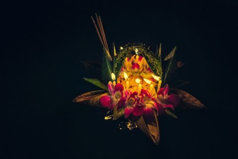 A krathong of folded leaves and flowers with lit candles and incense sticks, floating on dark water