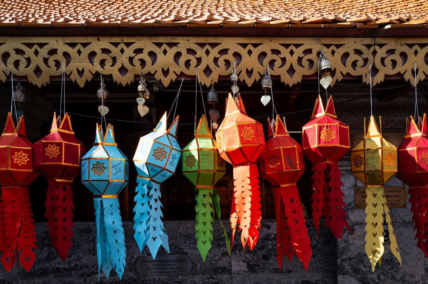 Red, orange, blue, green and yellow paper lanterns with long cut-paper tails, hanging in a row beneath a carved wooden eave, with small bells between them