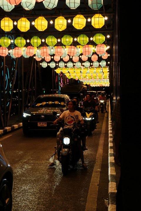 Rows of coloured paper lanterns hung over a bridge at night, with a car and motorbikes crossing beneath