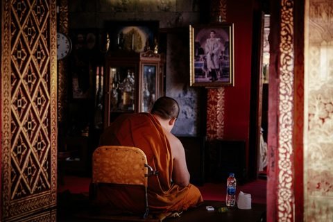 A monk in an orange robe, seen from behind, seated inside a temple hall between red-and-gold patterned pillars