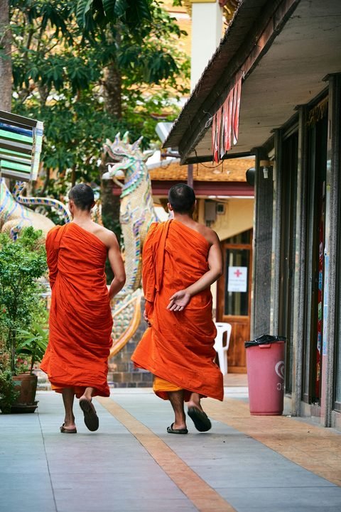 Two monks in orange robes walking along a pavement past a temple wall
