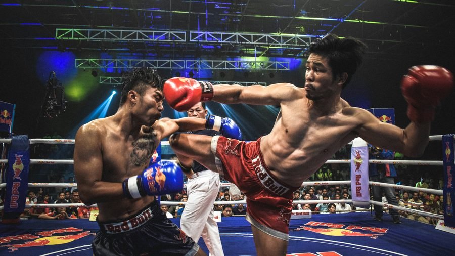 Two Muay Thai fighters mid-bout in a floodlit ring, one landing a high kick, with the referee and a full crowd behind them