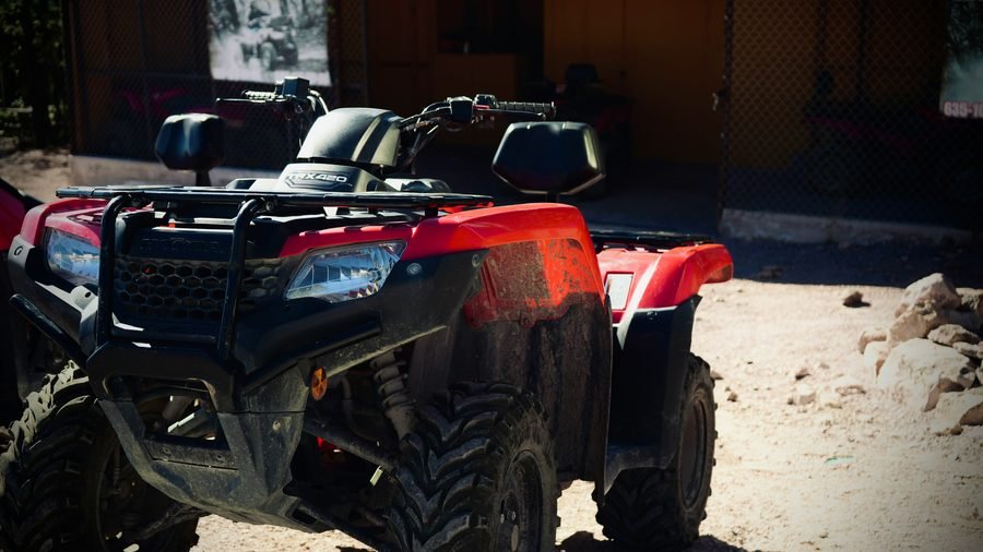 A red quad bike parked on a dirt track in front of the operator's shed, mud dried on its wheel arches