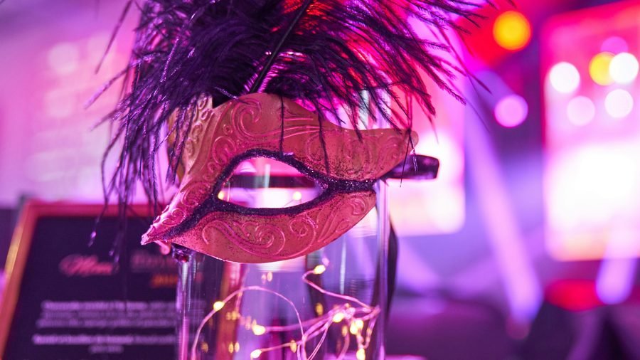 A feathered masquerade mask propped on a table under pink and purple stage lighting, the room behind it out of focus