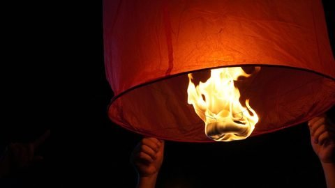 Close-up of the flame burning beneath an orange paper sky lantern, held up by hands in the dark