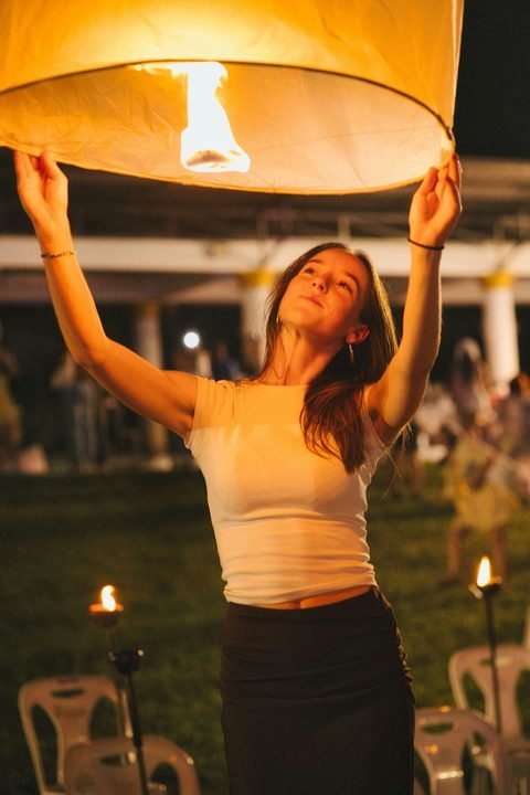 A person holding a lit sky lantern above their head on a lawn at night, with rows of chairs and small torches behind