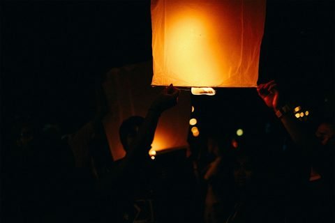 Raised hands holding a glowing paper sky lantern in the dark, with people in silhouette and a second lantern behind