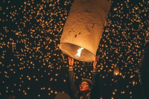 A person raising a lit sky lantern overhead, with hundreds of glowing lanterns filling the night sky behind